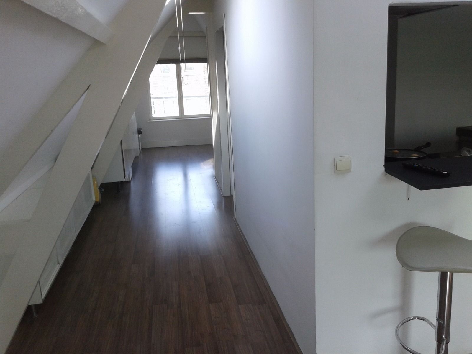 A narrow hallway with wooden flooring, a staircase on the left, and a black bar stool on the right.