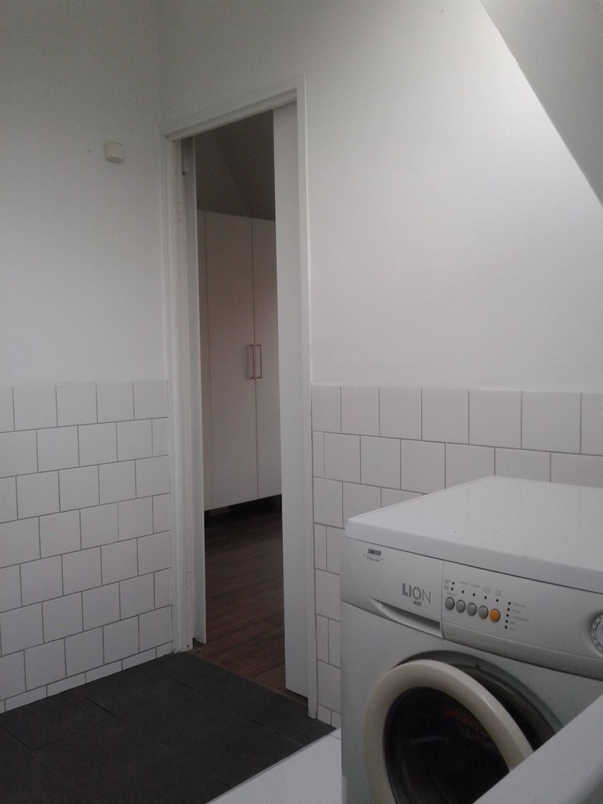 Une buanderie équipée d’un lave-linge à chargement frontal, avec des murs carrelés de couleur blanche et un plancher en bois.