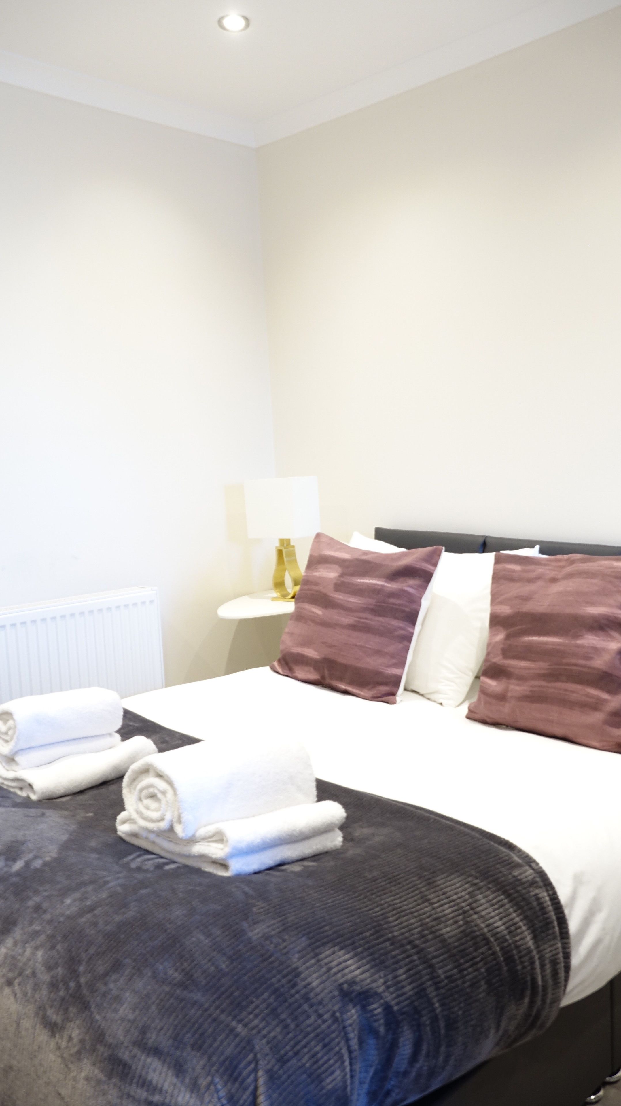 A hotel room with a neatly made bed, a bedside table with a lamp, and a radiator.