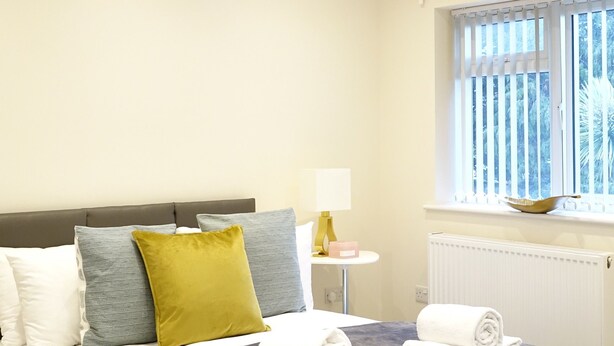 A modern bedroom with a bed, bedside table, lamp, and a window with blinds. - The Close Airport (Hayes)