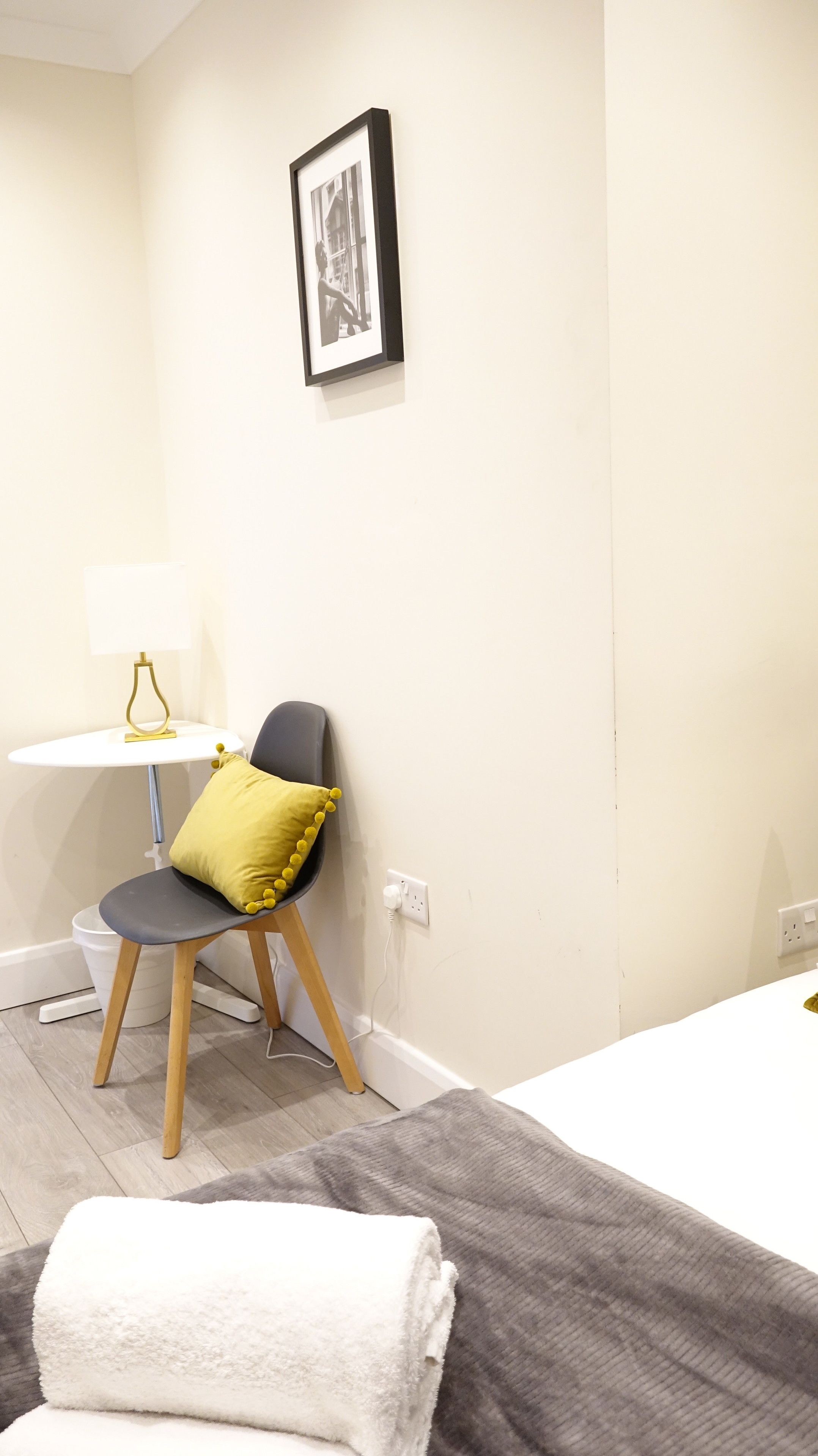 A hotel room with a bed, a chair with a yellow pillow, a small table with a lamp, and a framed picture on the wall.