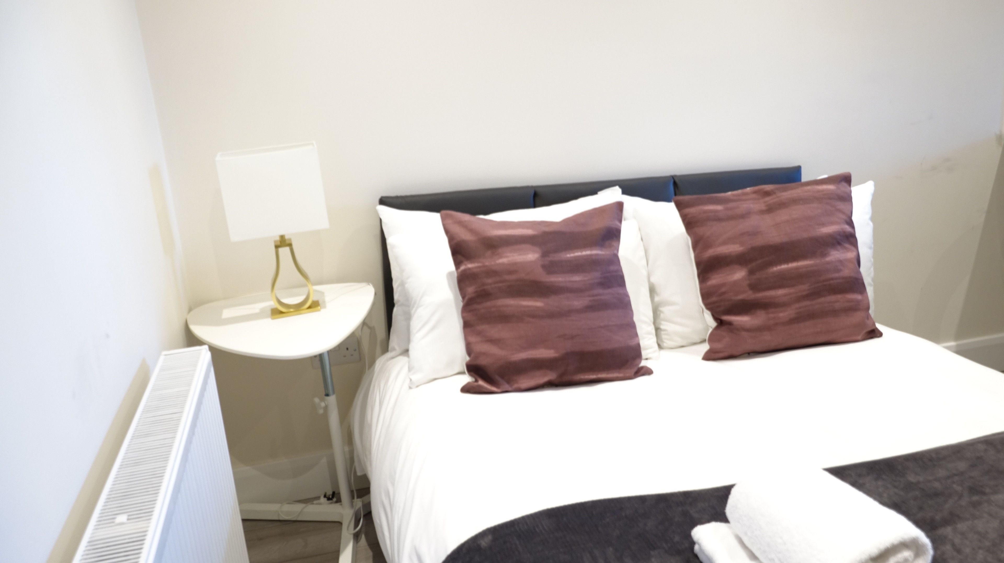 Une chambre d’hôtel avec un lit, du linge de lit blanc, deux oreillers marron, une table de chevet blanche avec une lampe et un radiateur blanc.