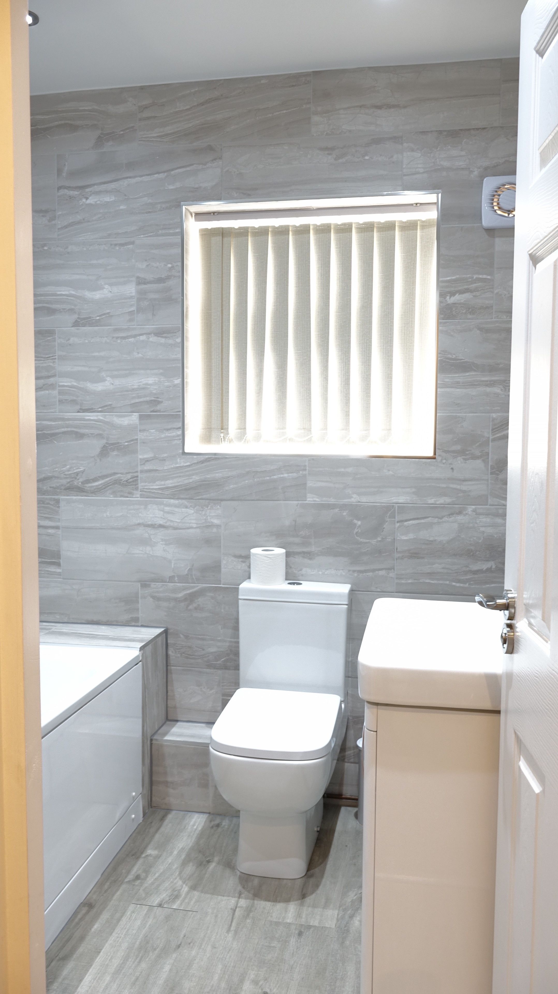 A modern bathroom with grey marble walls, a white toilet, and a window with vertical blinds.