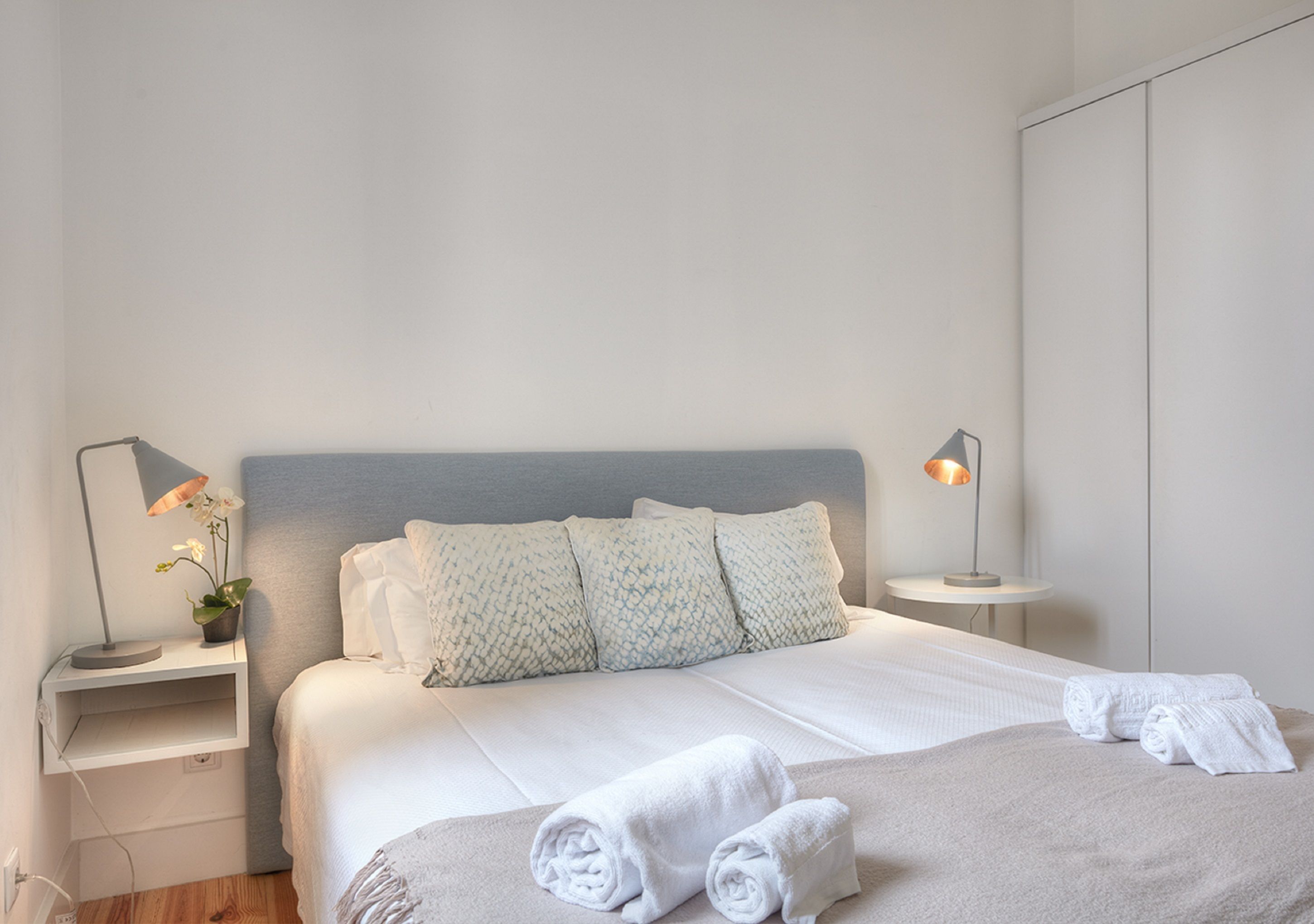 A neatly made bed with white linens and pillows, two bedside lamps, and rolled towels on the bed.