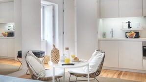 Une cuisine moderne avec des meubles blancs, un coin repas avec des chaises en osier et un espace pour le petit-déjeuner avec du jus d’orange et des viennoiseries.