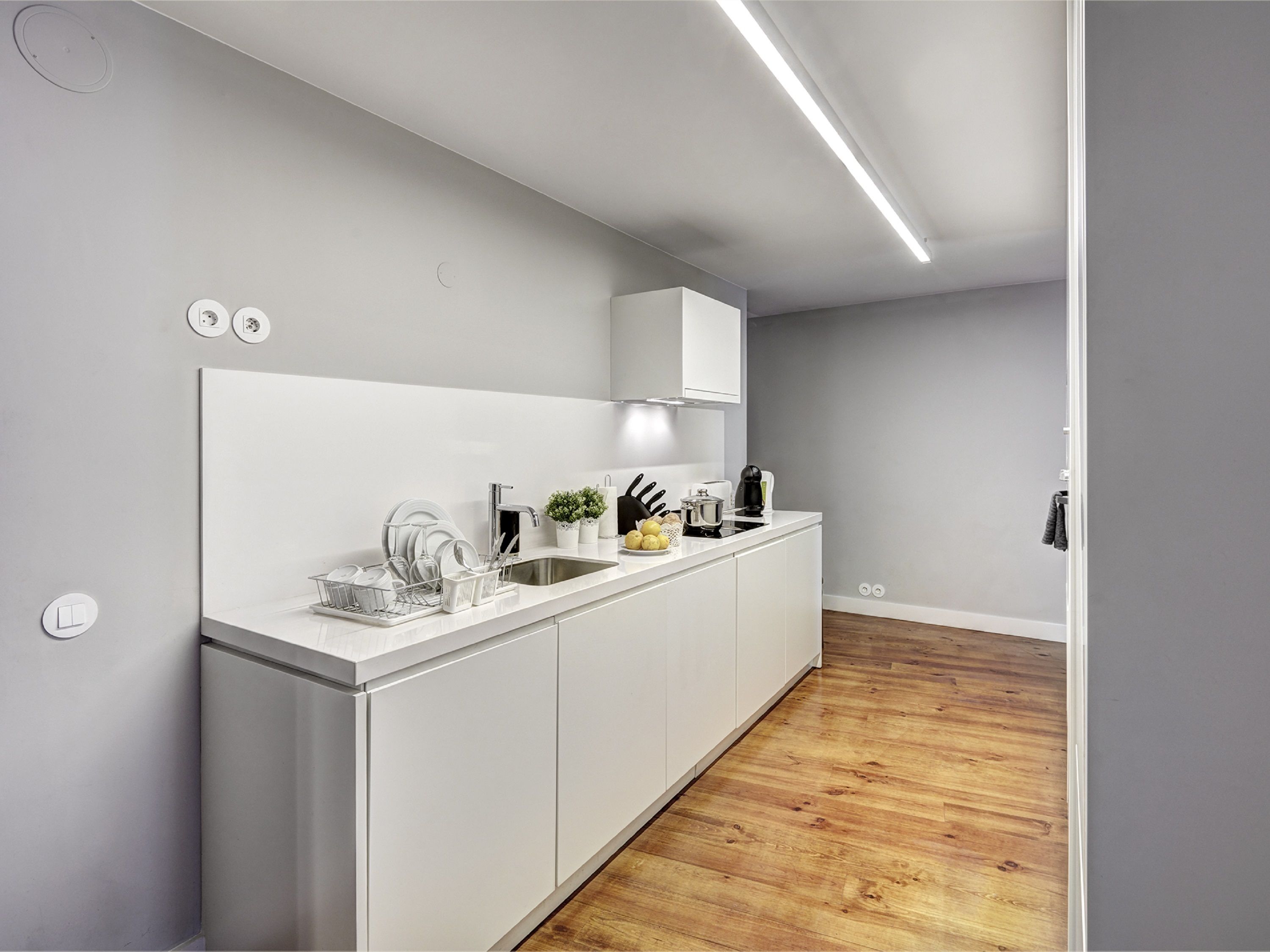 A modern kitchen with a white countertop, stainless steel sink, and wooden flooring.