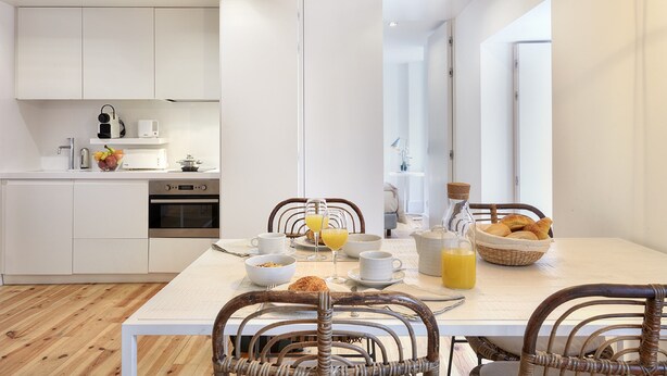 Une cuisine moderne avec des meubles blancs, une table à manger entourée de chaises en osier, et des aliments pour le petit-déjeuner, comme du jus d’orange et des croissants.