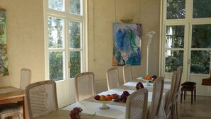 Een eetgedeelte met tafels voor vier personen, grote ramen en een schilderij aan de muur.