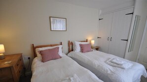 A hotel room with two single beds, a wooden nightstand with a lamp, a framed picture on the wall, and a white wardrobe. - Green Door Rose (Port Isaac)