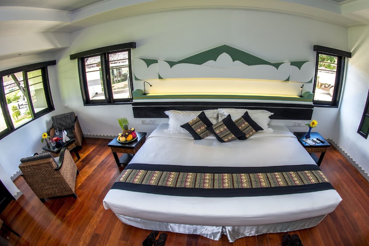 Uma cama grande com cabeceira branca e preta, duas mesas de cabeceira com abajures, uma pequena mesa com frutas e um vaso, e vista para o verde pelas janelas.