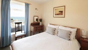 A bedroom with a bed, a desk, a chair, a window with curtains, a framed picture on the wall, and a nightstand. - Green Door - Upper Tregudda (Port Isaac)
