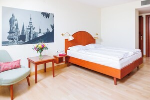 Una camera d'albergo con un letto di legno, un comodino, una sedia, un tavolino con un vaso di fiori e un quadro appeso al muro raffigurante un panorama urbano.