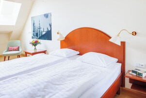 Un letto matrimoniale con lenzuola bianche, una testiera in legno, due lampade sui comodini, un comodino con dei libri e una poltrona con un cuscino rosa, oltre a un punto luce a parete.