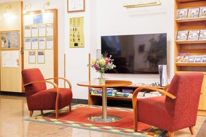 Uma área de recepção com duas poltronas vermelhas, uma mesa de madeira com um vaso de flores, uma TV de tela plana e uma estante com livros de viagem.