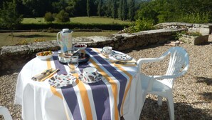 Une table à manger d’extérieur, dressée avec des assiettes, des bols, des tasses et des couverts, entourée d’une chaise blanche et d’un mur en pierre.