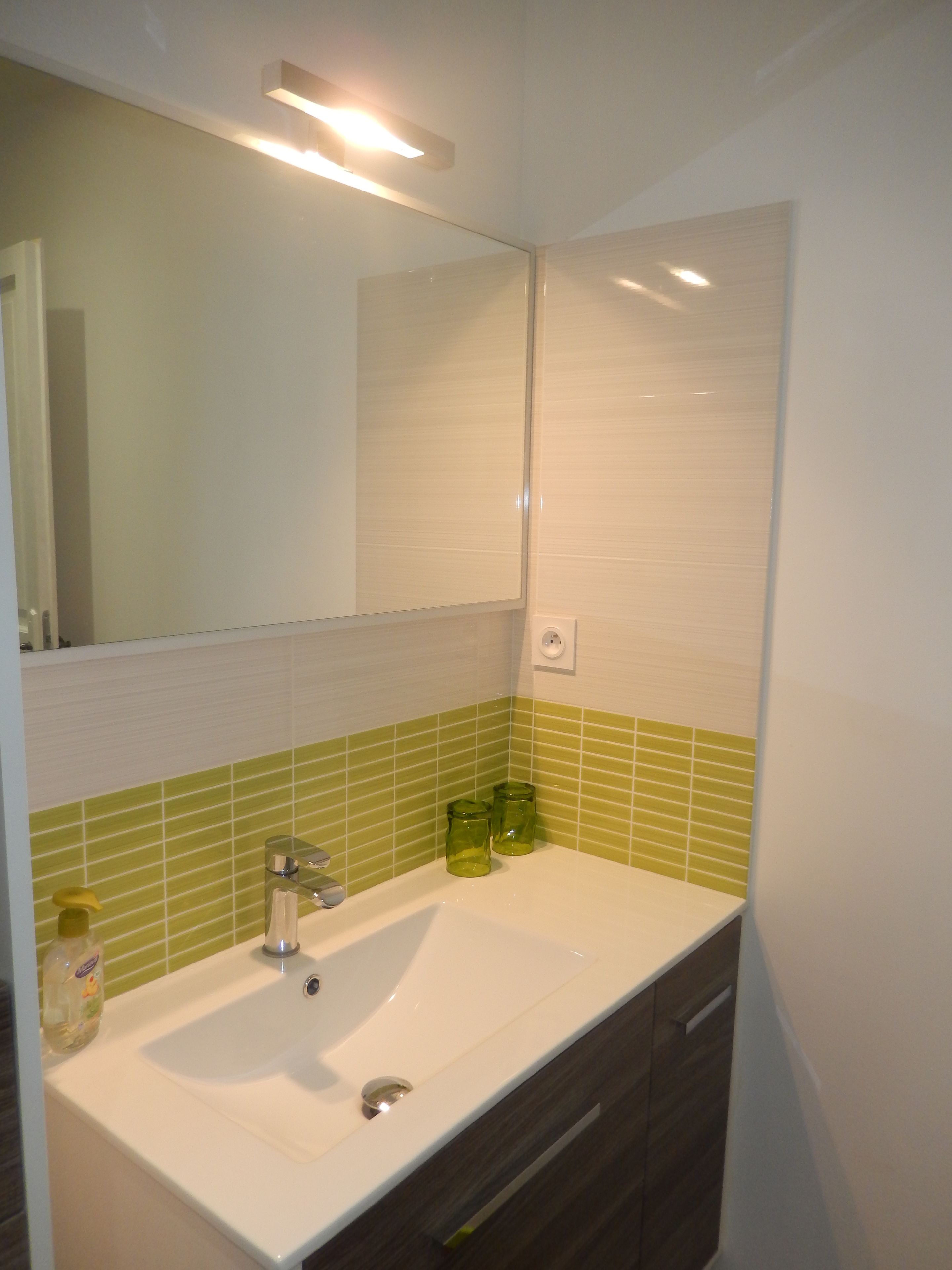A bathroom with a white sink, green-tiled walls, a mirror, and a light fixture above the mirror.