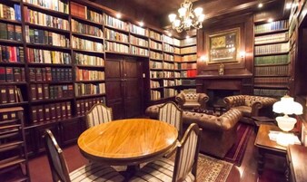 Uma biblioteca com estantes de madeira, uma mesa redonda de madeira, poltronas de couro e uma lareira.
