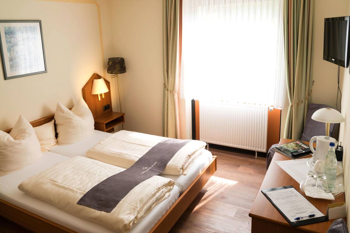 Ein Hotelzimmer mit zwei Betten, einem Schreibtisch mit Lampe, einem Fernseher und einem Fenster mit Vorhängen.