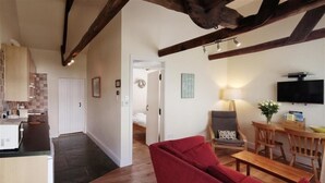 A cozy living area with a red sofa, wooden dining table, and a television. A kitchen with a microwave and a tiled backsplash is visible in the background. - Green Door - Thrift (Port Isaac)