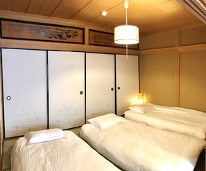 A traditional Japanese-style room with tatami flooring, a low ceiling, and sliding shoji doors. There are three beds with white bedding and a hanging light fixture.