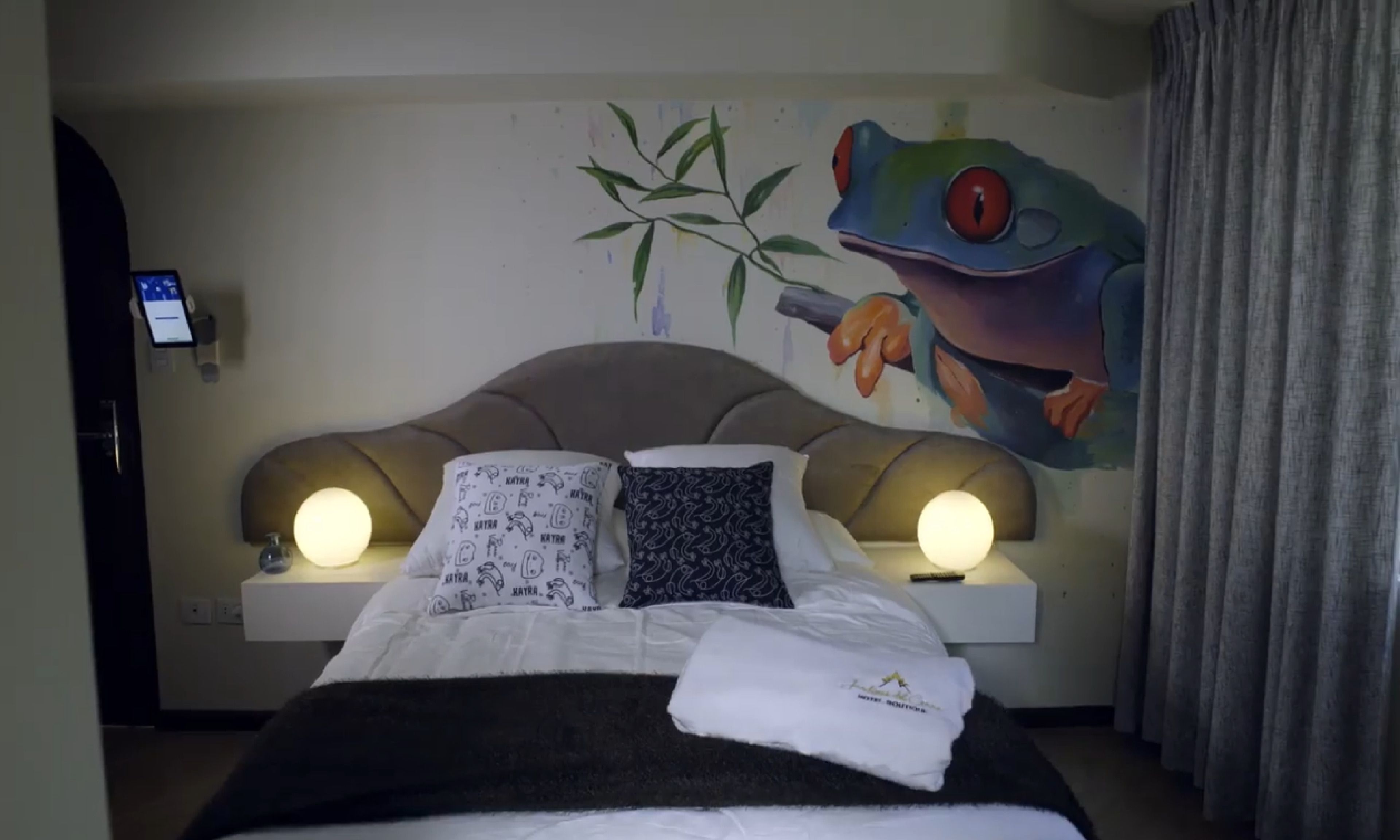 Habitación de hotel con cama, dos lámparas de noche, un mural de una rana y una estación de carga para teléfonos móviles.