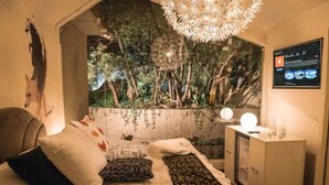 Un dormitorio con una cama, almohadas, una mesita de noche, una lámpara, un televisor montado en la pared y una pared decorada con plantas.