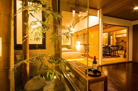A modern hotel room with a bed, a small table with a bottle and glasses, and a large window with bamboo plants.