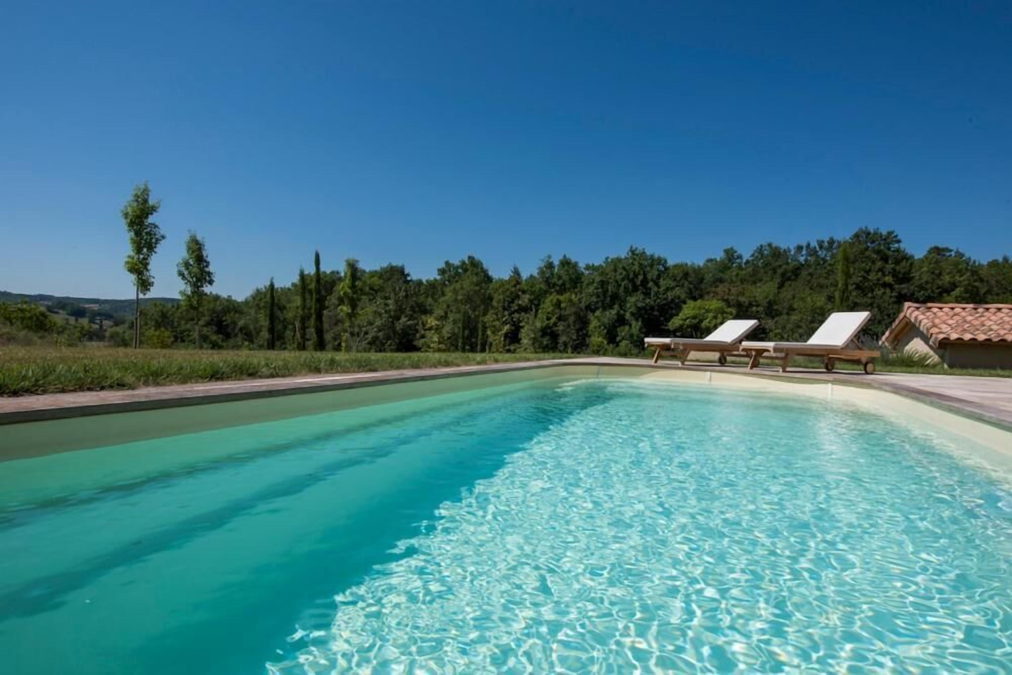 A swimming pool with clear water, surrounded by a landscaped area with trees and a building with a tiled roof in the background.