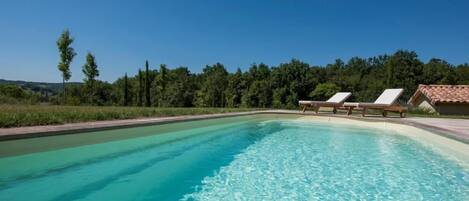 A swimming pool with clear water, surrounded by a landscaped area with trees and a building with a tiled roof in the background.