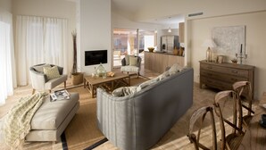 Een moderne woonkamer met een grijze hoekbank, een houten salontafel en een aan de muur bevestigde televisie. Er is een eethoek met een houten tafel en stoelen, en een keuken met een koelkast en magnetron.