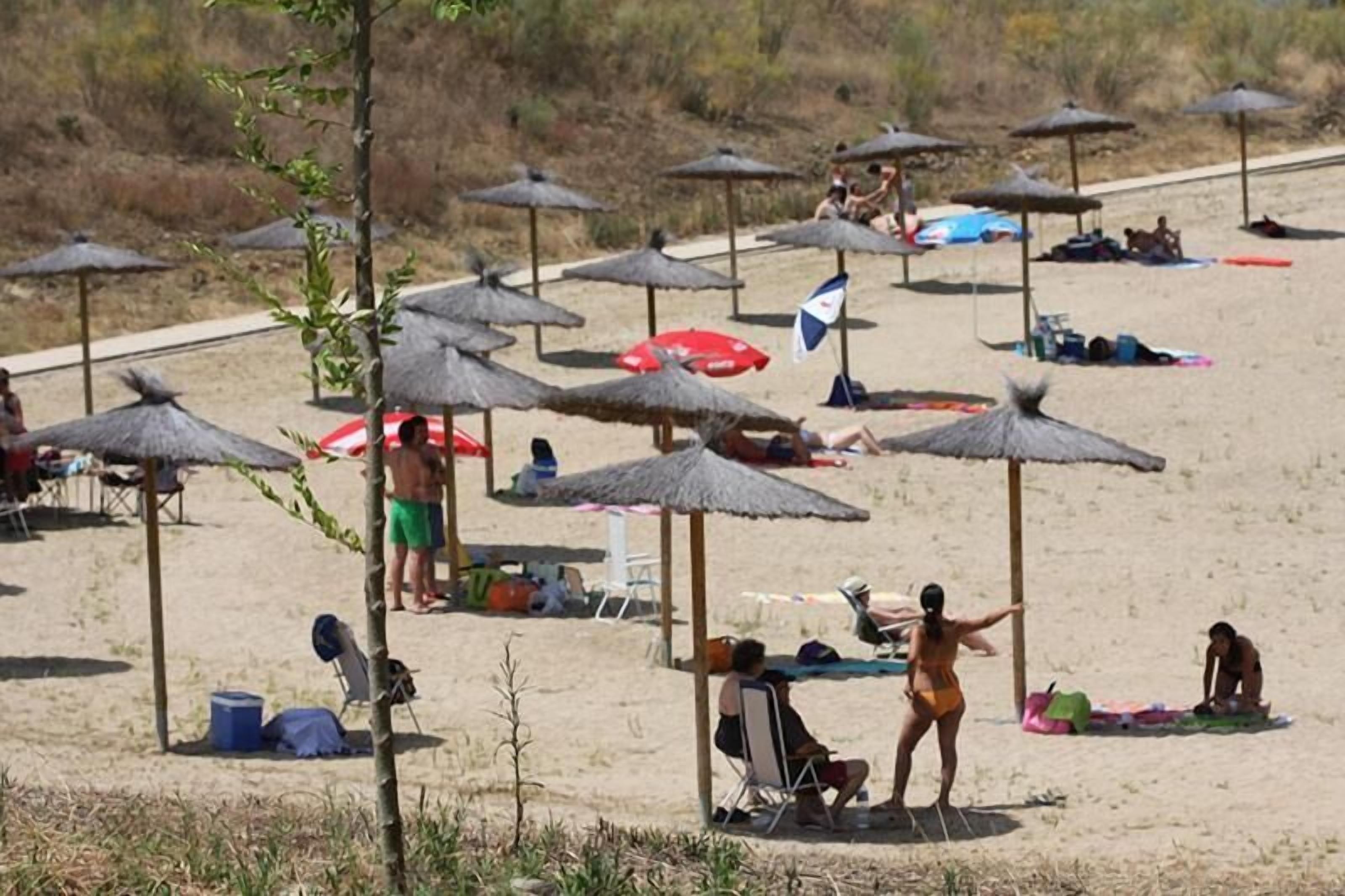 Una playa con sombrillas de paja y sillas, algunas personas tomando el sol y otras relajándose.