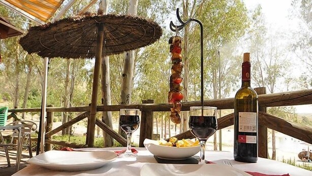 Un área de comedor al aire libre con una mesa preparada para dos personas, una botella de vino, un plato de comida y un pincho de carne colgando.