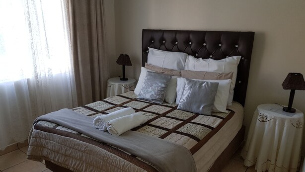 Cama bem arrumada com colcha, travesseiros e toalha dobrada. Há duas mesas de cabeceira com abajures.