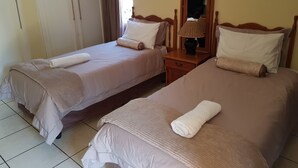 Duas camas de casal com roupa de cama bege e almofadas brancas, uma mesa de cabeceira de madeira com abajur e um espelho na parede.