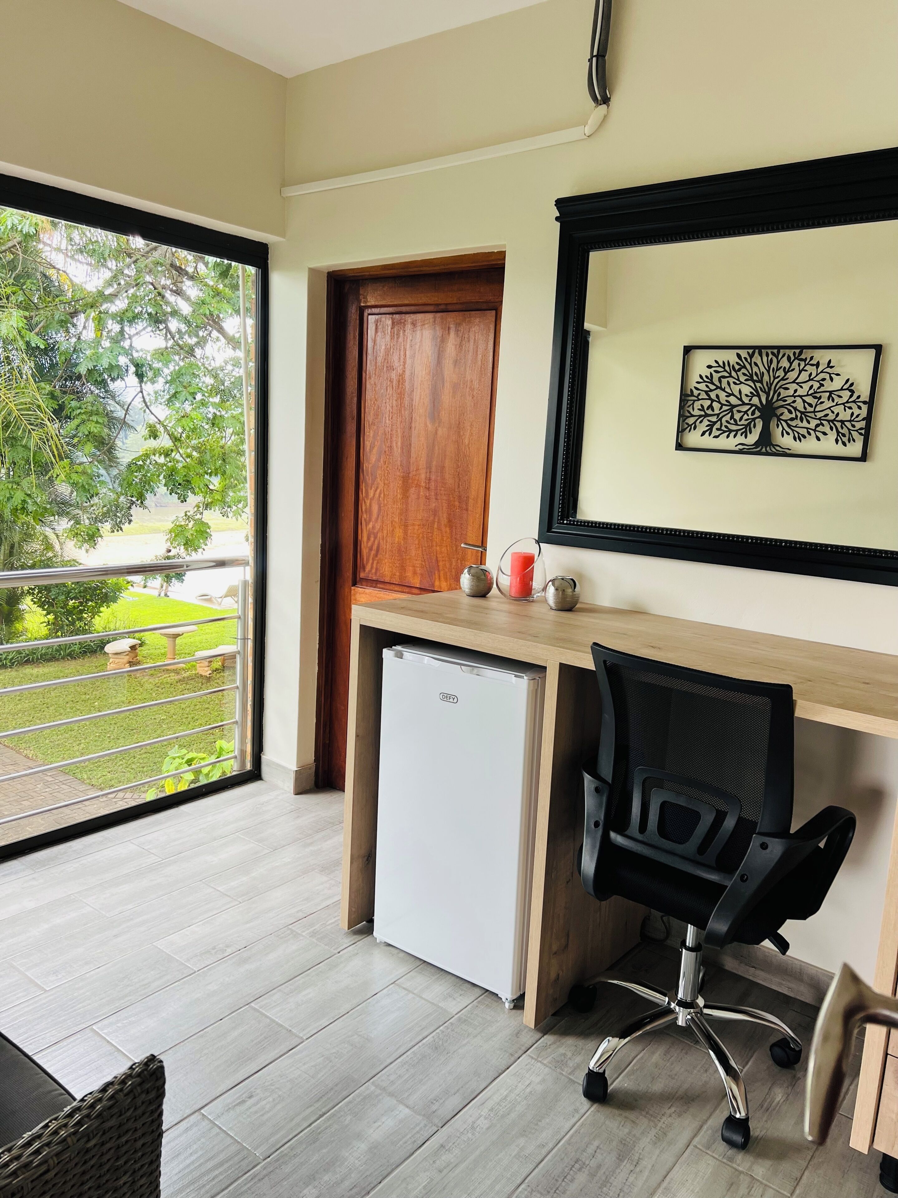A room with a wooden desk, a chair, a small refrigerator, a door, and a window with a view of greenery.