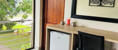 A room with a wooden desk, a chair, a small refrigerator, a door, and a window with a view of greenery.
