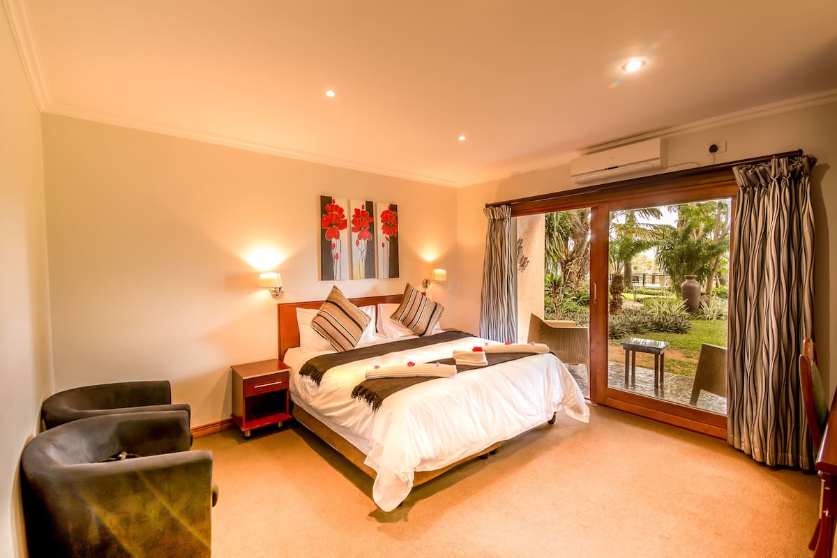 A bedroom with a bed, two bedside tables, a nightstand, a window with curtains, and a view of a garden.