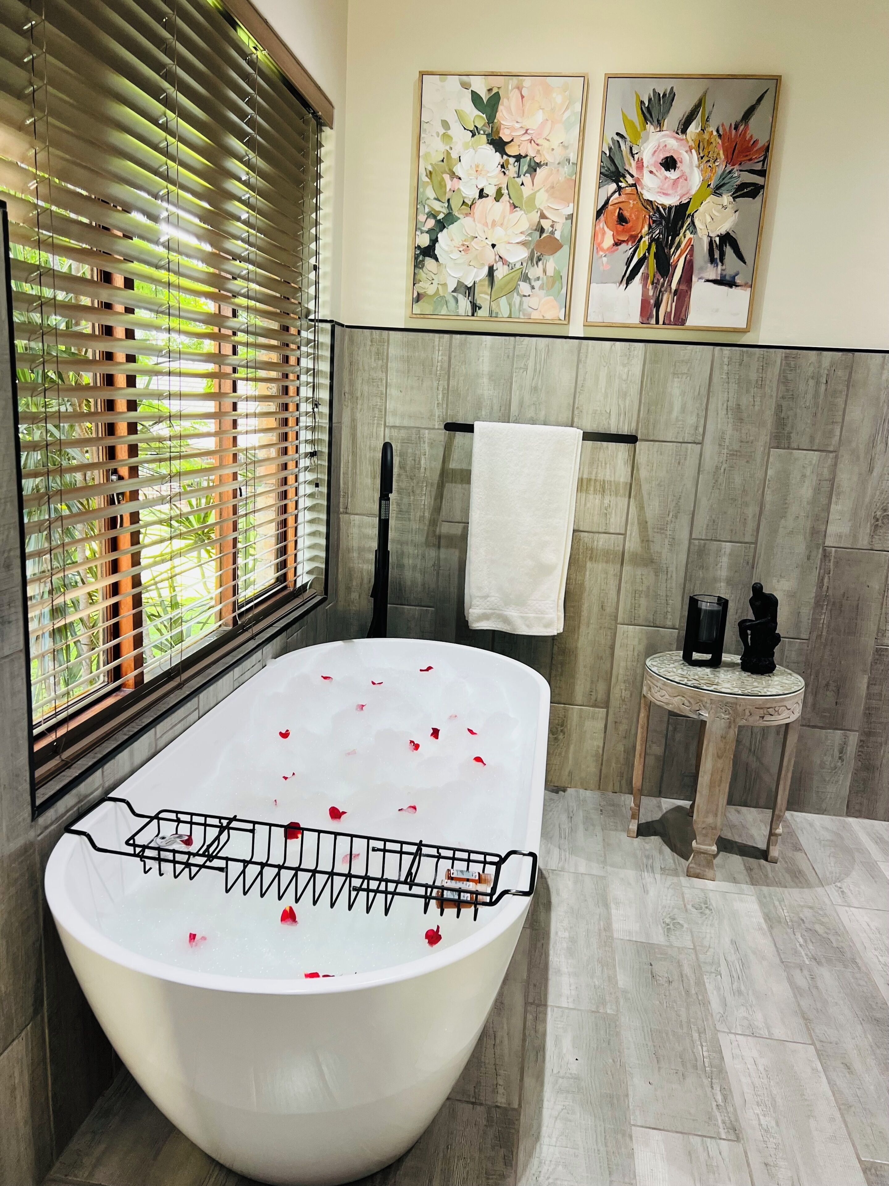 Een moderne badkamer met een vrijstaand bad, een raam met zonwering en twee florale schilderijen in lijsten aan de muur.