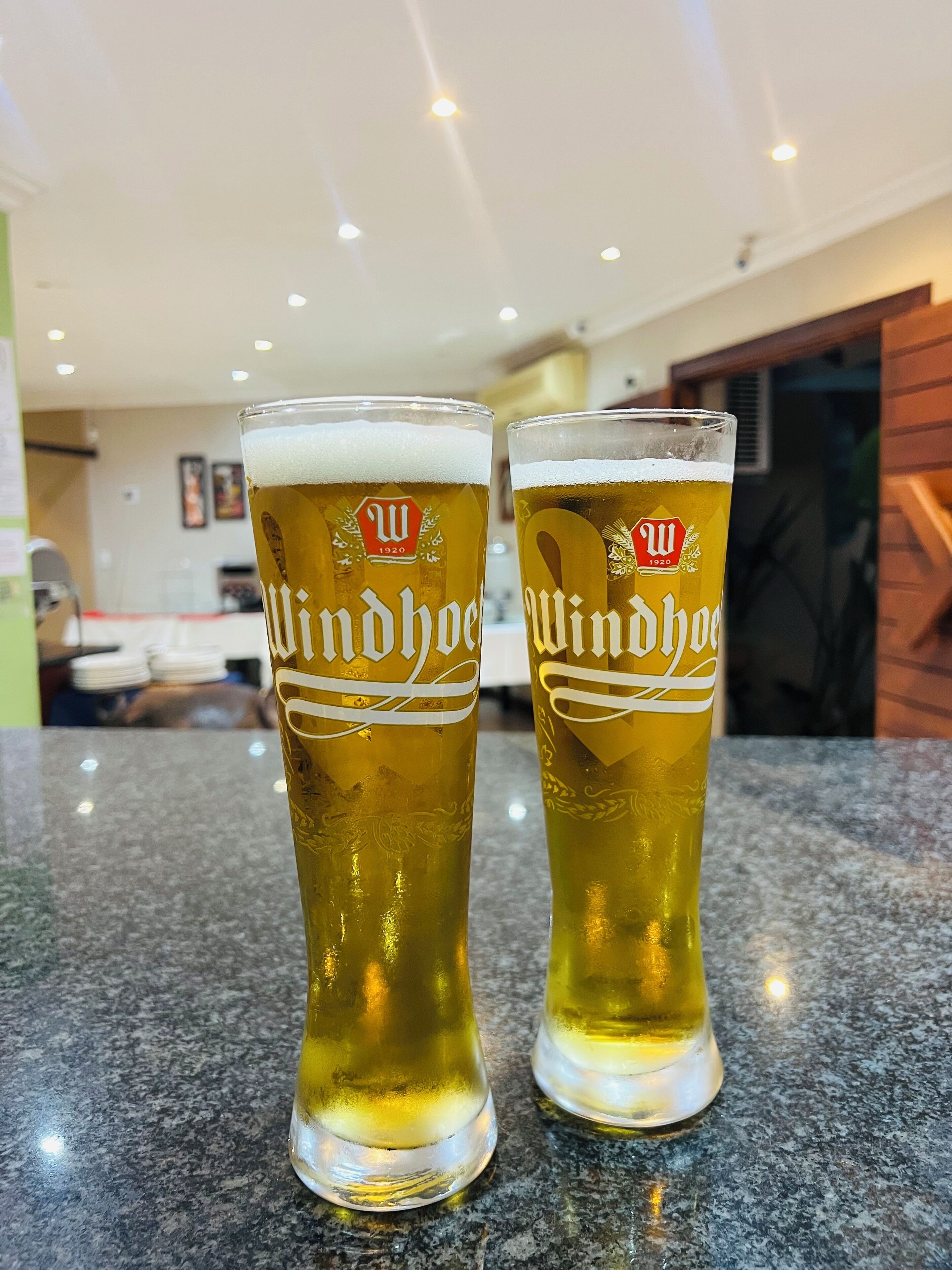 Duas taças altas de cerveja com a marca 'Windhoef' sobre uma bancada de granito em ambiente interno bem iluminado.