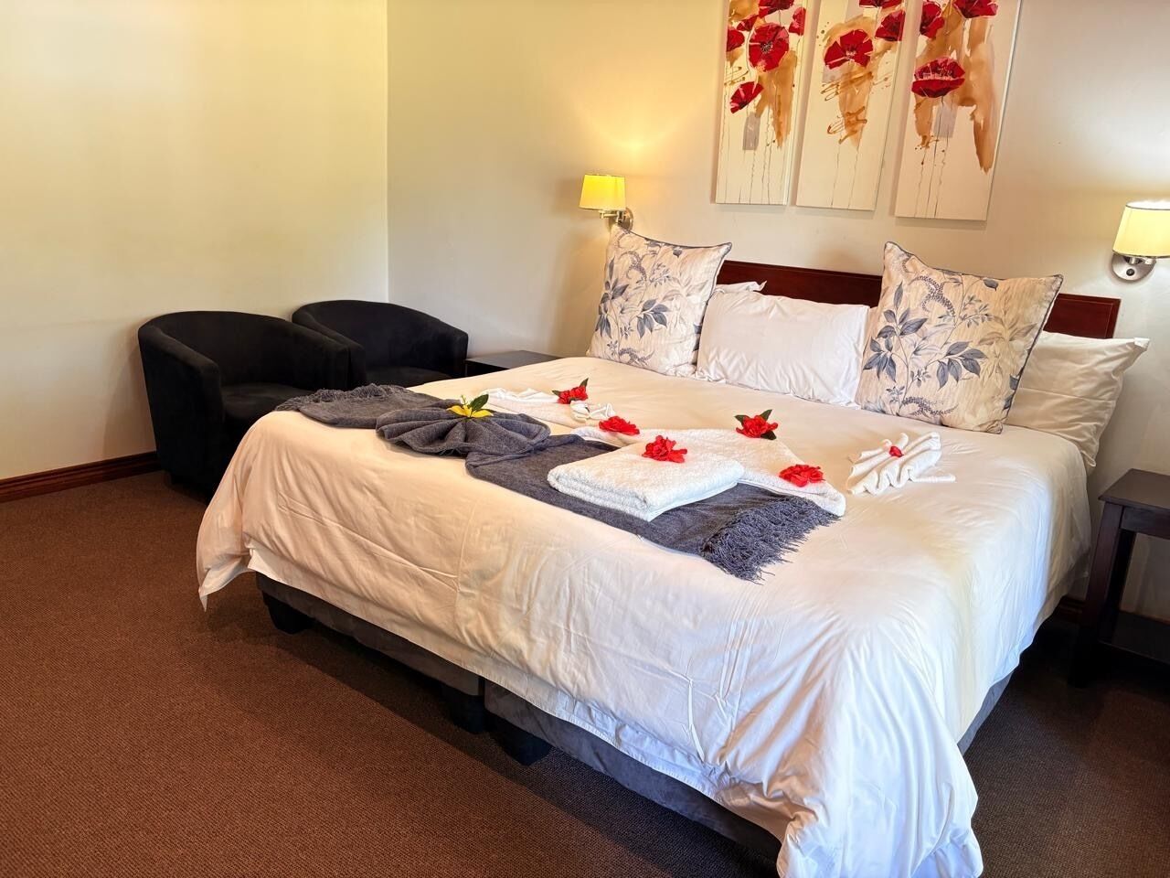 Una cama bien hecha con sábanas blancas, una manta gris y flores rojas. Hay dos lámparas de noche, un sillón oscuro y una mesita.