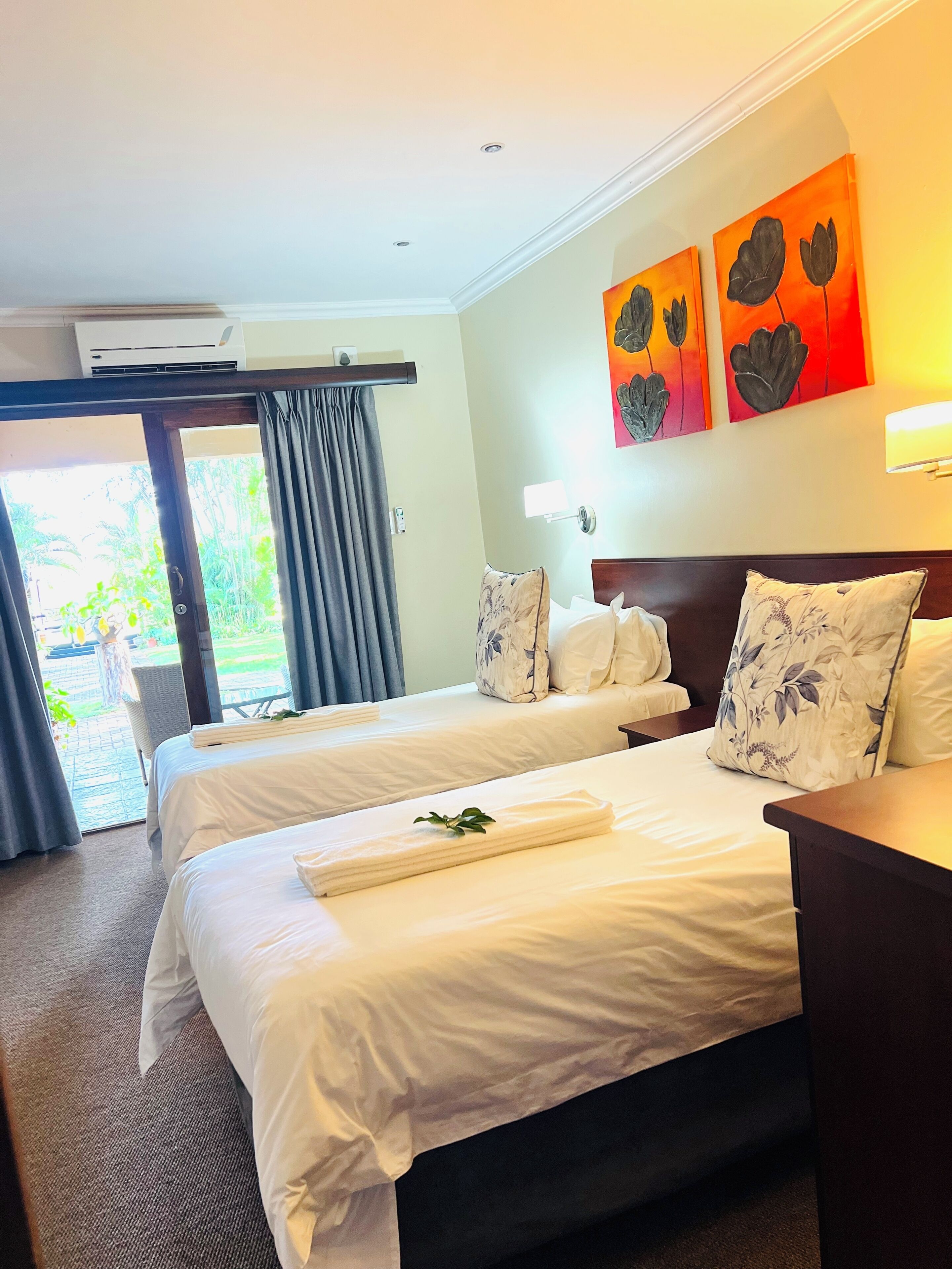 A hotel room with two beds, a large window with curtains, a wall-mounted air conditioner, and a painting above the beds.