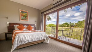A bedroom with a large bed, a nightstand, a painting on the wall, and a balcony with a view of greenery.