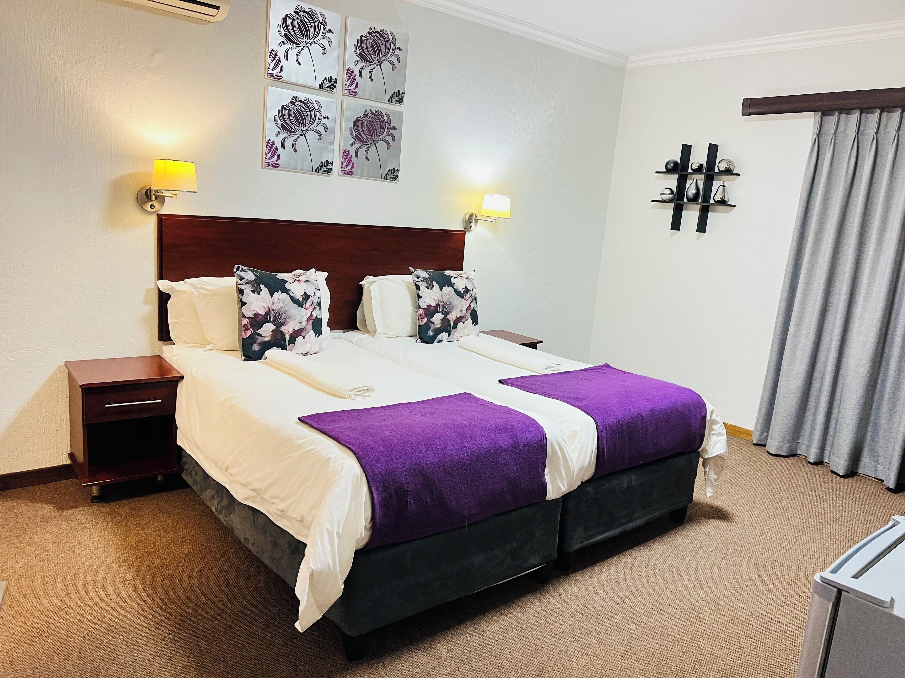 A hotel room with a large bed, two nightstands, a headboard, a wall-mounted air conditioner, a wall-mounted shelf with decorative items, and a window with curtains.