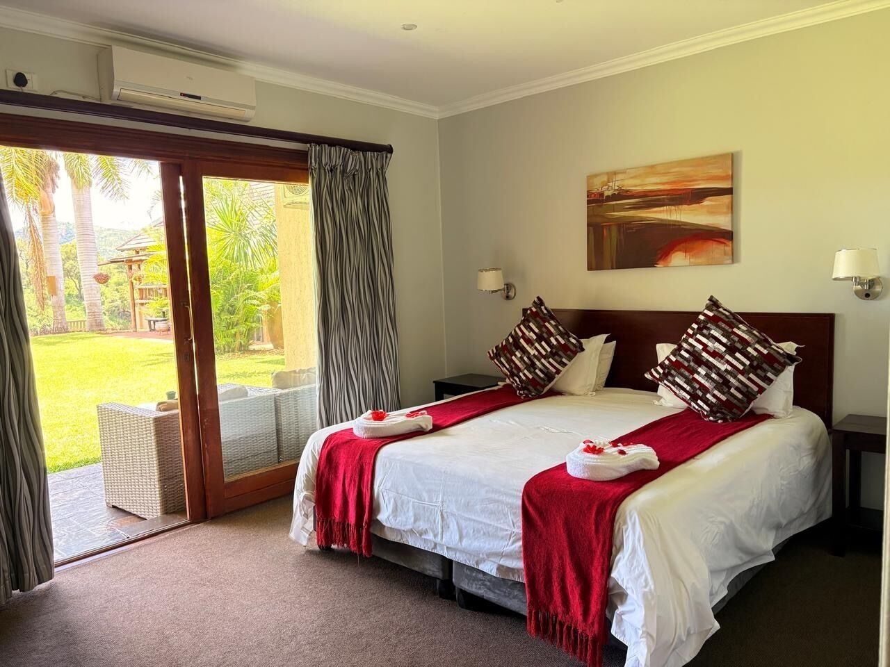 A hotel room with a large bed, a sliding glass door to a garden, and a painting on the wall.