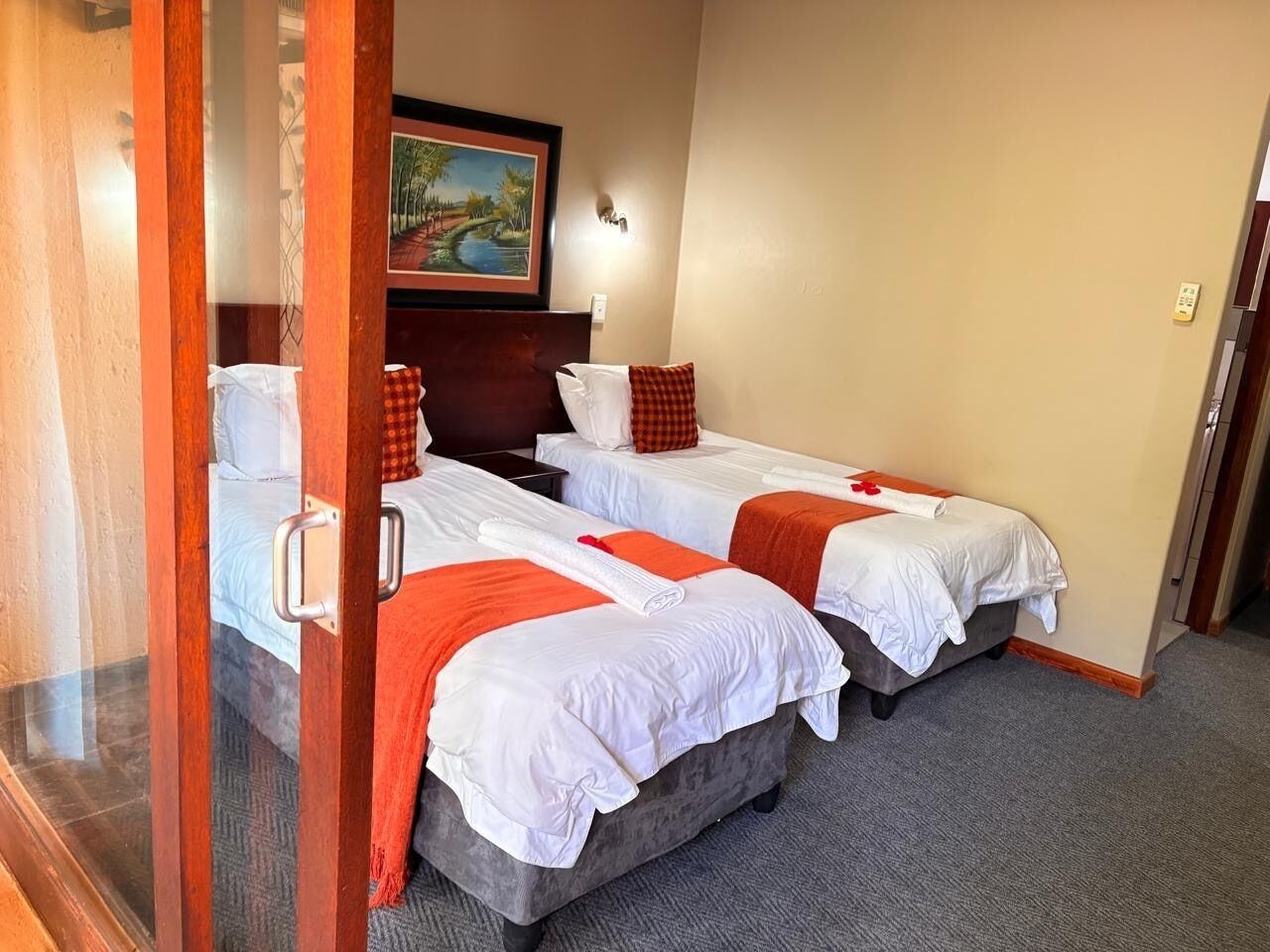 Ein Hotelzimmer mit drei Einzelbetten, jedes mit weißer Bettwäsche und orangefarbenen Akzenten. Über den Betten hängt ein Gemälde und dazwischen steht ein kleiner Tisch.