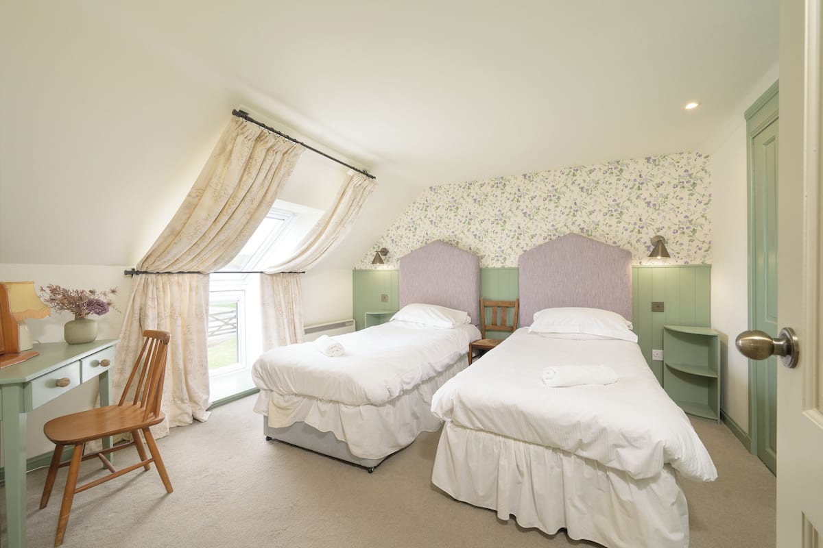 A bedroom with two beds, a desk, a chair, a window with curtains, and a floral wallpaper.