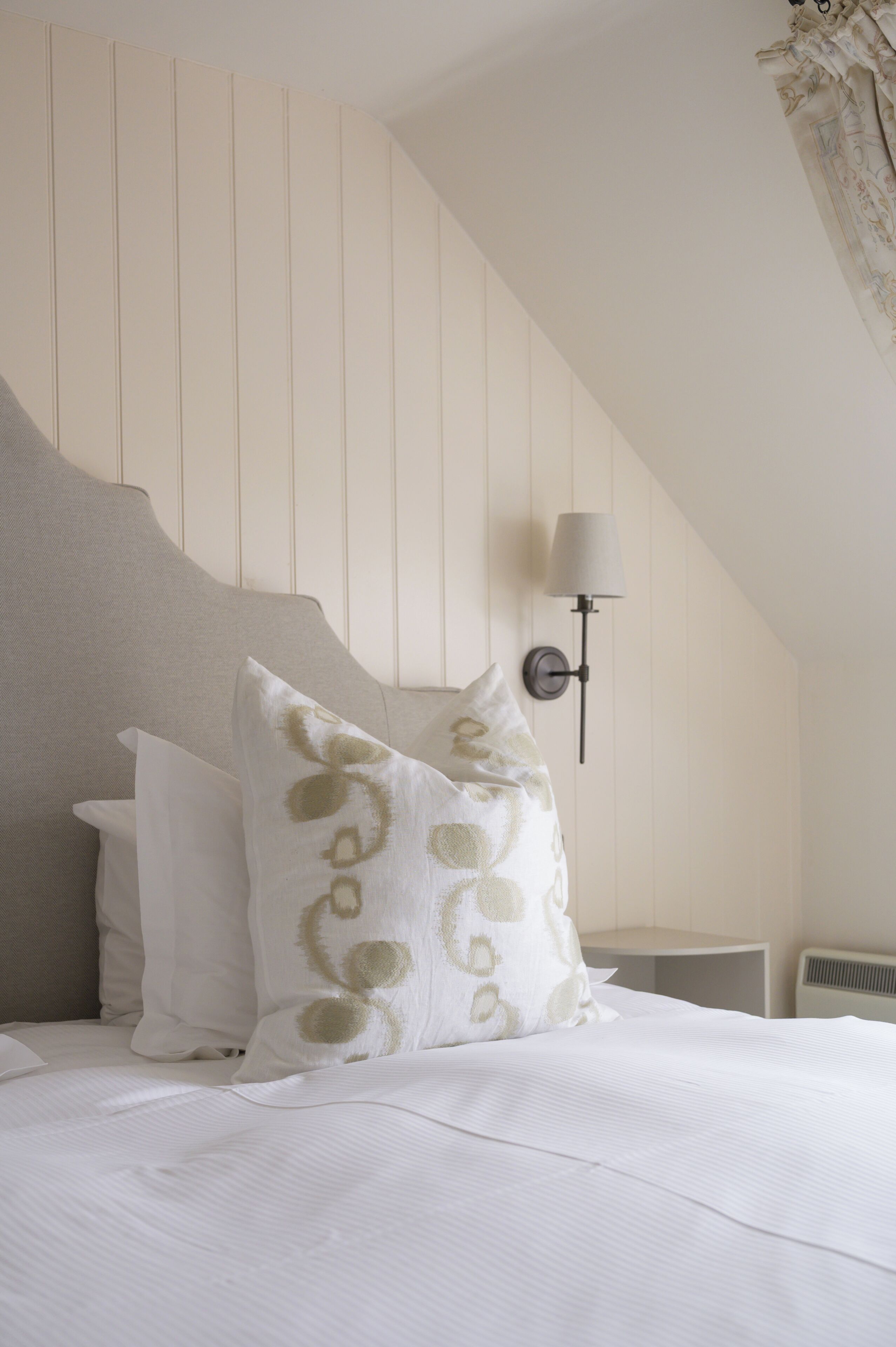 A bedroom with a bed, pillows, a wall-mounted lamp, and a sloped ceiling.