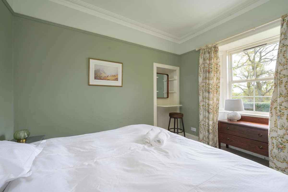 A neatly made bed in a room with a green wall, a framed picture, a small stool, and a window with curtains.