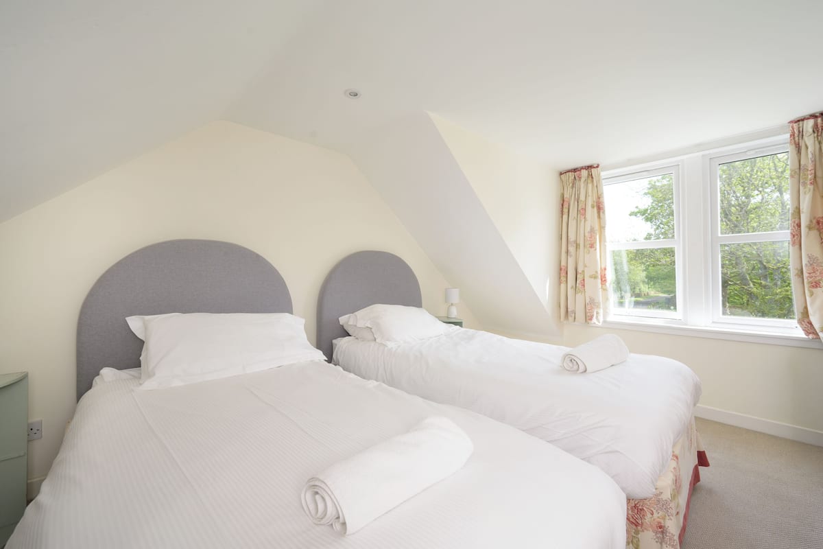A bedroom with two beds, a window with curtains, and a nightstand with a lamp.