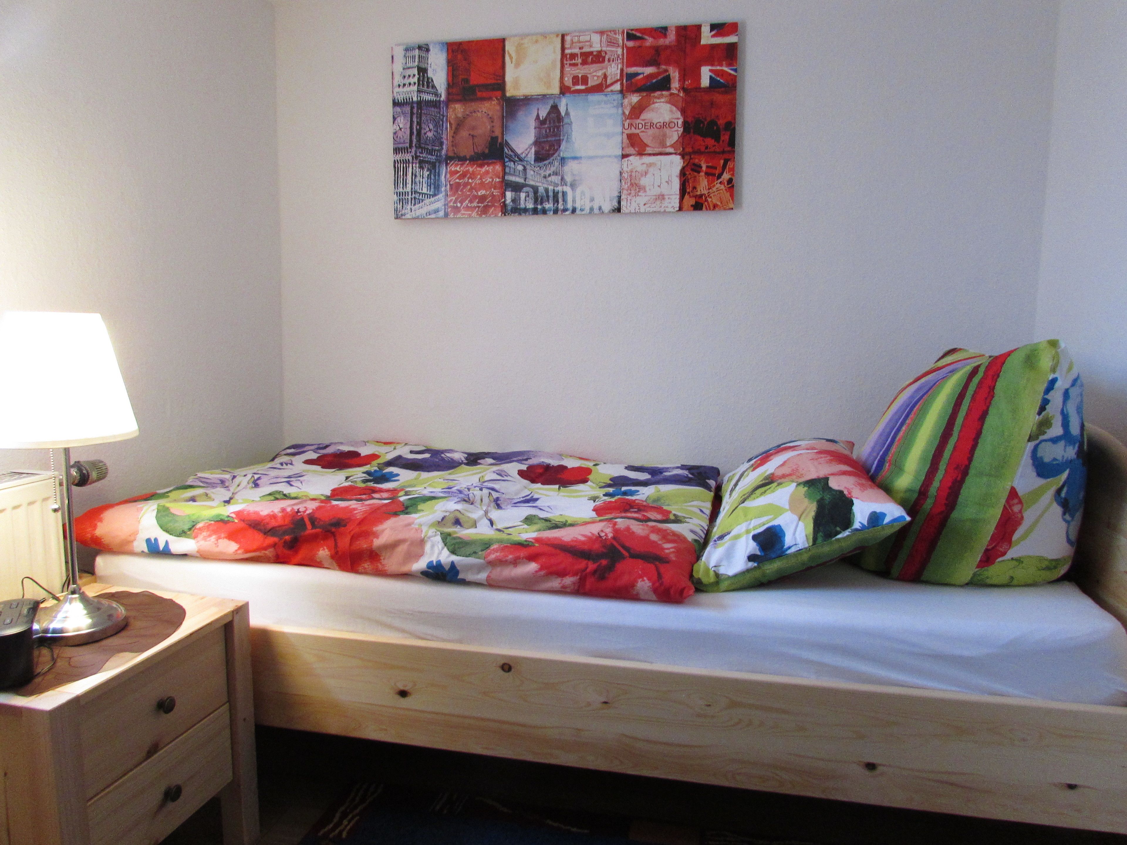 Une petite chambre avec un lit simple, un cadre en bois, une table de chevet avec une lampe et une couvre-lit à motifs floraux aux couleurs vives.